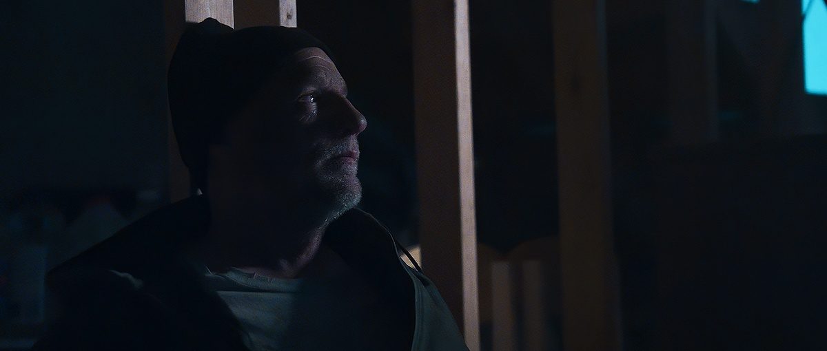 Mac, played by Daniel J. Lennox, standing in a dim room.